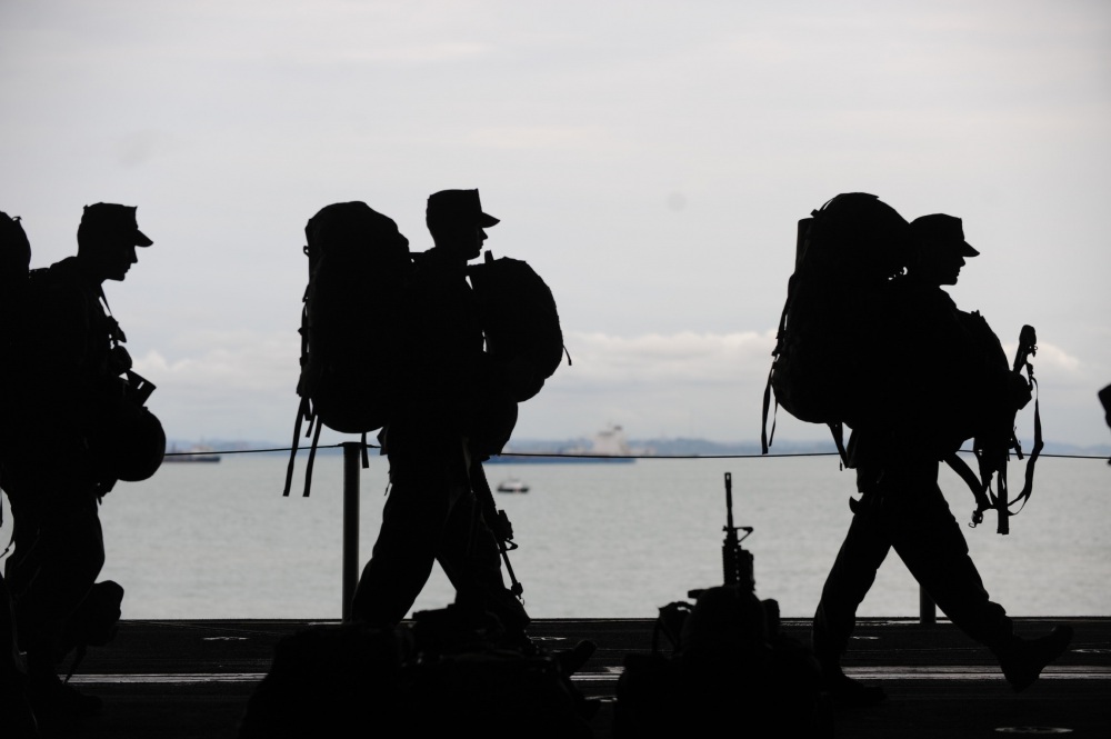 3 soldiers carrying large backpacks are silhouetted with a harbor in the background.